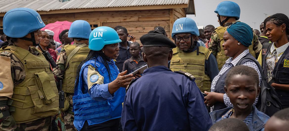 The image captures a group of people, including several UN peacekeepers wearing distinctive blue helmets and vests, in a crowded, outdoor setting. The peacekeepers, mostly wearing camouflage uniforms, stand among civilians and engage in conversation. The background features a wooden structure with a corrugated metal roof. Several individuals are visible in the crowd, some wearing casual clothing, while others appear to be in uniforms. The scene seems busy, with people interacting closely, and a mixture of attentive and engaged expressions can be seen throughout the group.