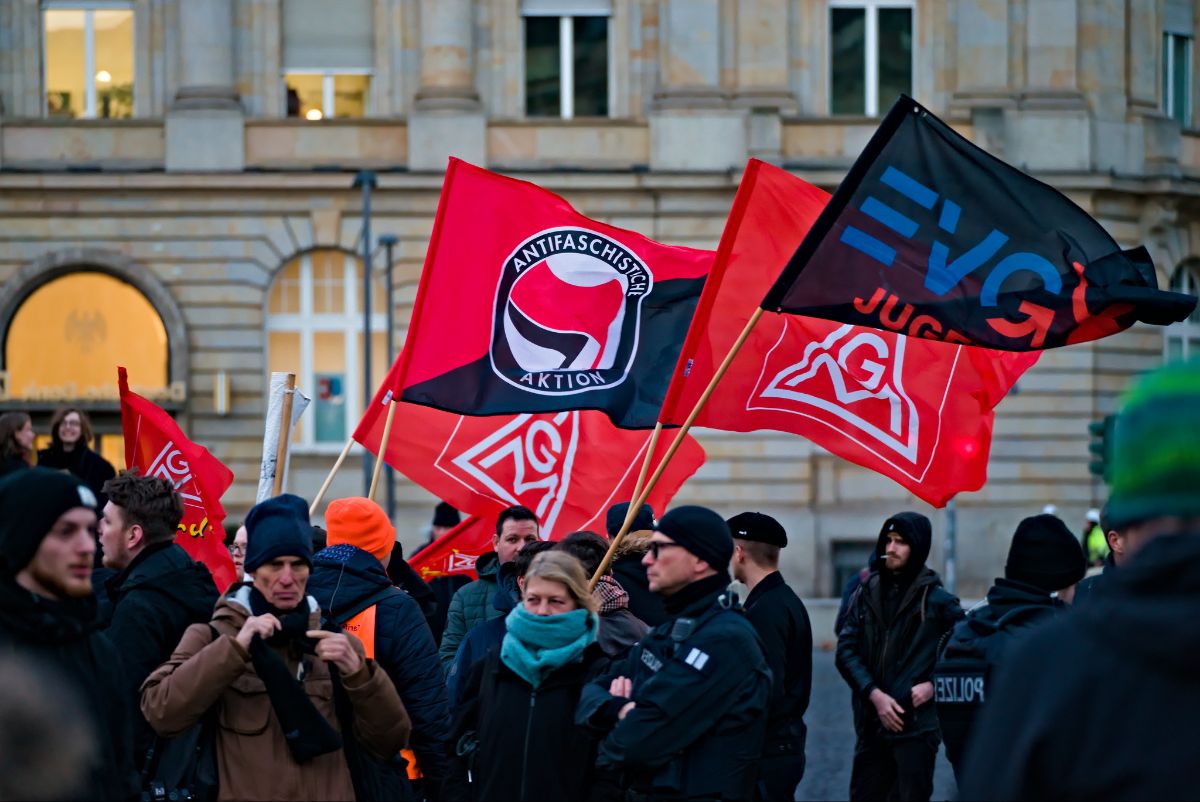 The image depicts a gathering with multiple individuals participating in a demonstration or protest. Several people are standing closely together in a public space, many of whom are dressed in winter clothing like jackets, hats, and scarves, indicating cold weather. Prominently displayed are several brightly colored flags, mostly red and black, held by some participants. The flags have various logos and text, including an emblem with the words “ANTIFASCHISTISCHE AKTION” surrounding a circle with stylized shapes inside. The background shows an architectural façade of a building with large windows, enhancing the urban setting. The atmosphere appears engaged, with participants facing in different directions.