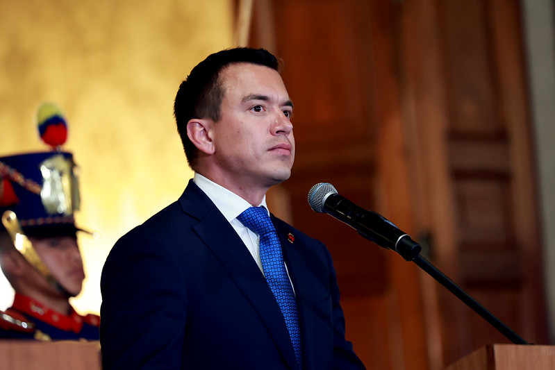 The image features a man in formal attire standing behind a podium and speaking into a microphone. He is wearing a dark blue suit jacket, a white shirt, and a cobalt blue tie with a subtle pattern. The background features wooden paneling and a blurred figure dressed in a ceremonial military uniform on the left, with a hat adorned with a plume and colorful details. The setting appears to be formal and possibly governmental or official in nature.