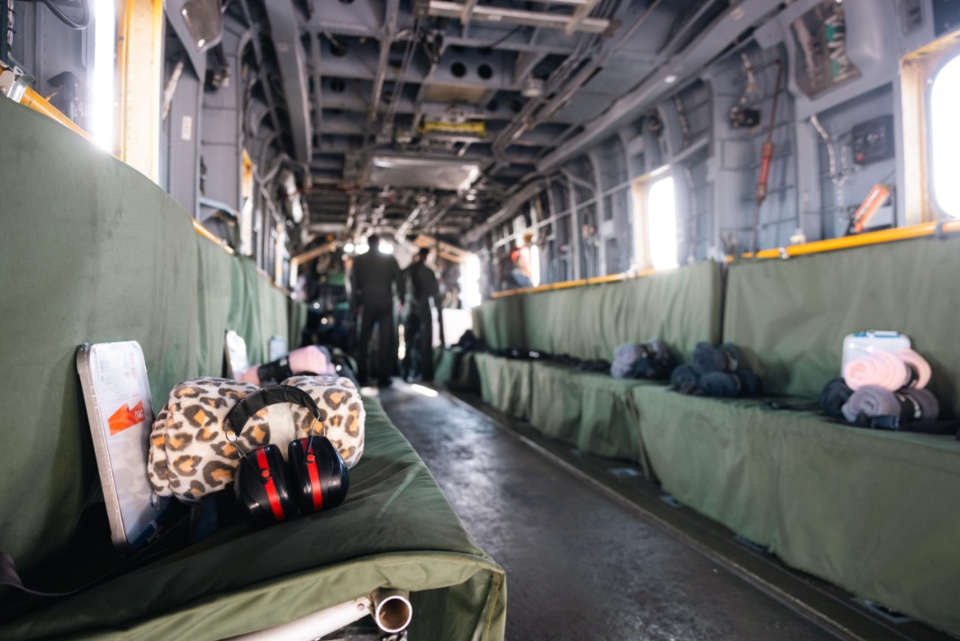 The image portrays the interior of a large military or cargo aircraft. The perspective is from the front towards the back of the aircraft, where two figures stand silhouetted. The cabin is spacious, with a high ceiling and lined with green fabric-covered benches on both sides. On the left bench, a leopard-print neck pillow with black and red headphones rests towards the foreground. Additional travel essentials are visible on the benches, including a pink neck pillow, gray rolled blankets, and packaged items. The lighting is relatively bright, coming from windows along the aircraft's sides, illuminating the rugged metallic interior structure with exposed beams and wires.