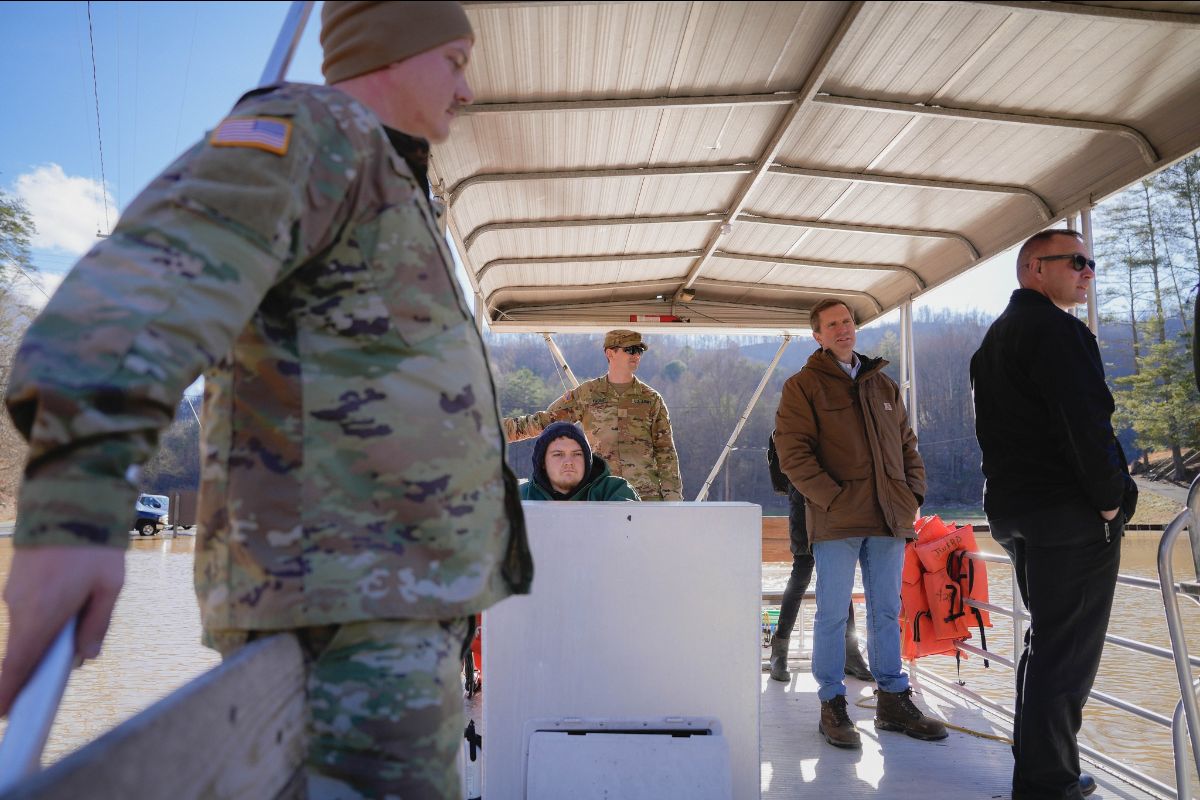 The image depicts five individuals standing under a metal canopy on a pontoon boat. In the foreground on the left, a person in camouflage military uniform and a brown beanie is holding onto the boat's railing. In the middle of the image is a person sitting at a helm in a dark hoodie. Behind them, another individual in a camouflage uniform stands, wearing a cap and sunglasses. To the right, two men in civilian clothing are standing, one in a brown jacket with blue jeans, the other in a black jacket and sunglasses. Orange life jackets are attached to the railing. The boat is on a body of water, and the background shows a forested hillside against a clear blue sky.