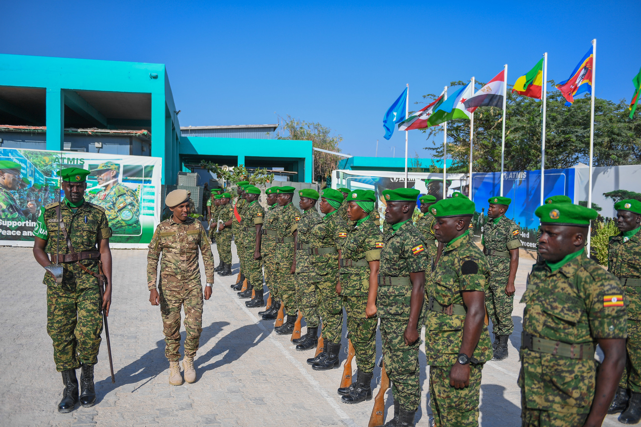 The image shows a formation of soldiers in camouflage uniforms and green berets standing in a line. A person in a different camouflage uniform, wearing a beige beret, walks in front of the soldiers. The setting appears to be an outdoor area with a clear blue sky and a large turquoise building in the background. Several flags are mounted on white poles, fluttering in a row beside the building. In the background, there is a large banner featuring imagery of soldiers and text that is partially visible.