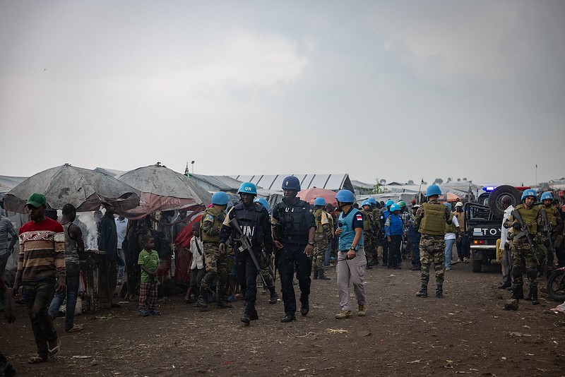 The image depicts a group of peacekeepers wearing blue helmets and uniforms marked with "UN" patrolling a busy outdoor market area. The market consists of several makeshift stalls covered by tarps and populated by civilians. Behind the group of peacekeepers, there are military vehicles and personnel. The ground is muddy, and the sky is overcast, suggesting possible rain. The atmosphere is bustling with several individuals either engaged with the stalls or walking through the area.