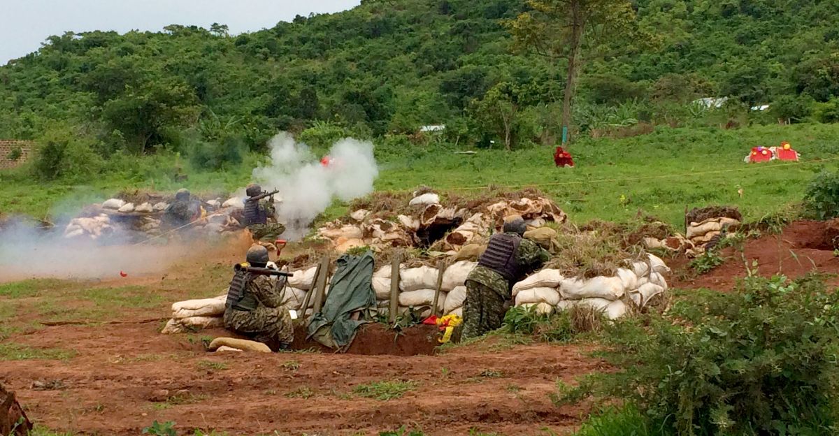 The image depicts a scene of military personnel engaged in a training exercise. Several soldiers, wearing camouflage uniforms and protective gear, are positioned behind a makeshift barricade constructed of sandbags and earth. Smoke billows from the area, indicating gunfire or simulated explosions. The soldiers are crouched in various positions, aiming firearms towards a target. The setting is an open, grassy field with a backdrop of lush green hills and scattered trees.