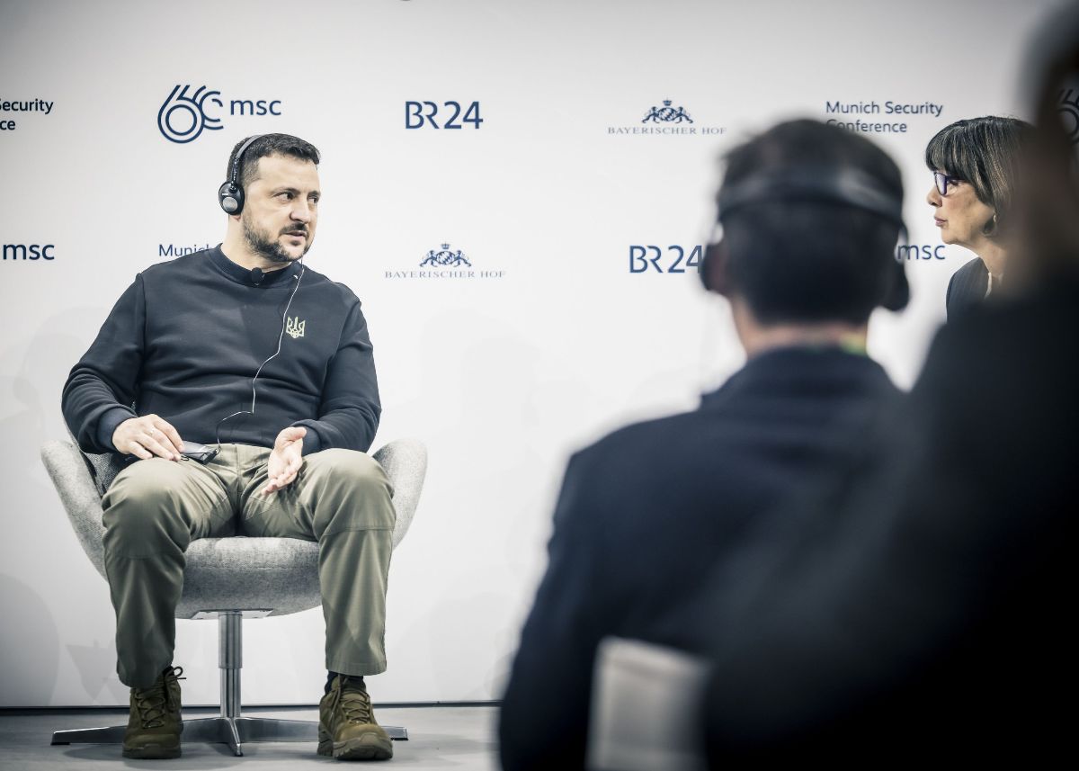 The image shows a man sitting on a light gray chair, engaged in a conversation. He has short dark hair and is wearing headphones, a black sweatshirt with an emblem, olive green pants, and brown shoes. He is gesturing with his hands. Opposite him, a woman with short dark hair and glasses is visible, seated and facing him. In the foreground, the back of another person's head is visible, slightly out of focus, suggesting the presence of an audience. The background is white with logos and text, indicating a formal setting related to a conference or event. The lighting is soft, creating a professional atmosphere.