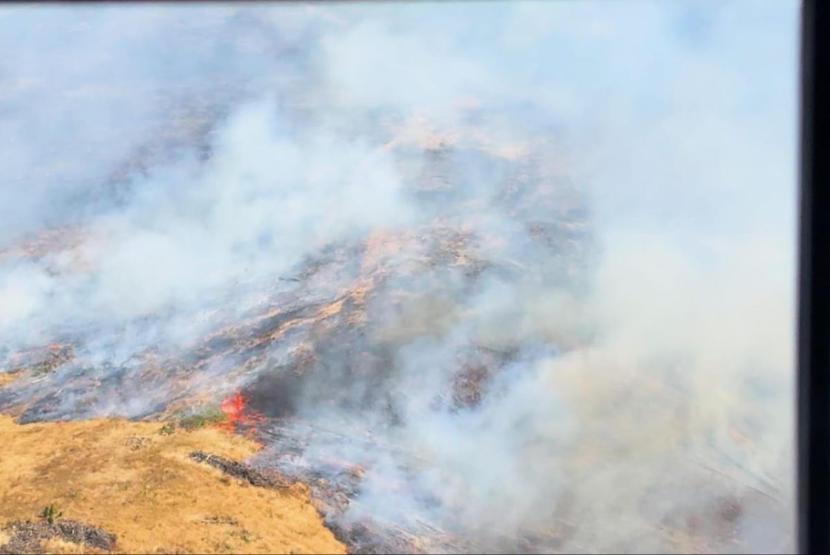 The image shows an aerial view of a landscape engulfed in smoke, likely from a fire. The scene is covered with thick, white smoke that obscures much of the ground. Beneath the smoke, patches of burned or burning land are visible, with darker areas indicating more intense burning. In the bottom left, there is a section of dry, yellowish grass or vegetation. A small, bright red flame is visible near this area, suggesting active fire. The overall atmosphere is hazy due to the pervasive smoke.