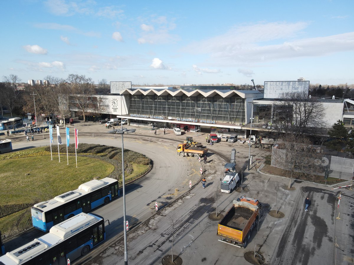 The image depicts a large, modernist-style building with a distinctive zigzag roof pattern in the foreground. The facade features a large glass wall with angular sections, bordered by white, concrete-like panels. In front of the building, there is a construction area with several vehicles, including a truck and a small utility vehicle. Workers can be seen near the construction area, with some equipment visible. In the foreground, a circular road surrounds a small green area with grass and a few landscaped sections. Several flagpoles with various flags are positioned near the road. Blue and white public buses are parked on the road, with trees and more urban structures in the background. The sky is clear with few clouds, suggesting a sunny day.