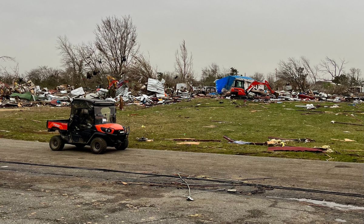 The image depicts a scene of devastation likely caused by a tornado. In the foreground, a small orange utility vehicle with a covered cabin is parked on a paved road. The background is filled with scattered debris, including twisted metal sheets and broken wooden structures, which are strewn across a grassy area. Among the rubble, a red excavator with a blue tarp is visible, suggesting ongoing cleanup efforts. Several leafless trees stand amidst the wreckage, and the overcast sky adds a somber tone to the scene.