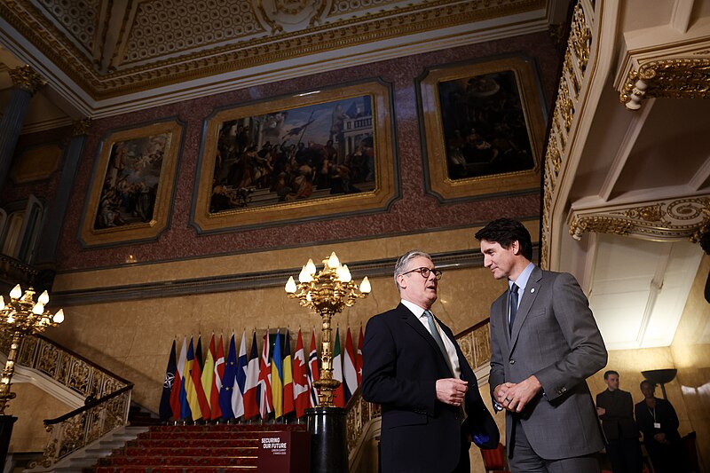 The image shows two individuals in a grand, ornately decorated hall. The setting features a staircase adorned with intricate metal railings and an array of international flags lined up along the red-carpeted stairway. Above them, the walls display large, framed classical paintings set within a lavishly decorated interior with gold accents and detailed moldings. Two ornate lamps with multiple bulbs illuminate the area. The atmosphere is formal and the overall color palette includes rich golds, warm beiges, and deep reds.