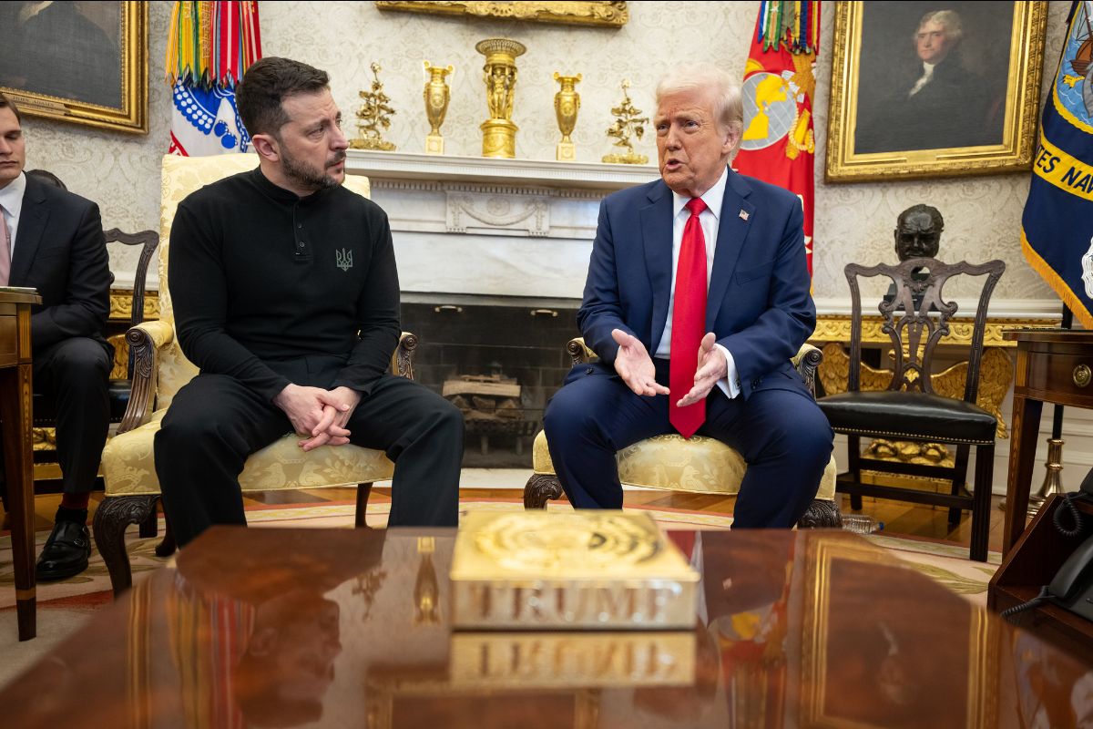 On the left is a man in black fatigues with a black logo over the heart. On the right is an older man with a blue suit and red tie. They sit in an ornate room