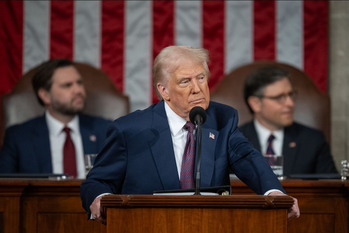 The image shows a man in a suit standing at a podium with a microphone. He has light hair and is wearing a dark blue suit with a white shirt and patterned tie. On the lapel of his suit is a small American flag pin. Behind him, two men are seated, both wearing suits and ties. They appear slightly blurred, with one man on the left donning a red tie and the other on the right with a blue tie. The background features a large American flag hung vertically along the wall.