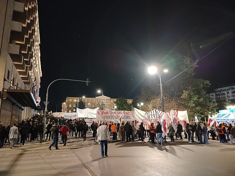 The image depicts a nighttime protest scene in an urban setting. A large crowd of people is gathered in the street, many holding up banners with text. The street is well-lit by high streetlights, creating bright spots against the dark night sky. On the left side, a multi-story building with a series of balconies lines the street. A light post curves over the crowd near the building. In the distance, a notable building with illuminated windows and a symmetrical façade stands, likely a government or significant public building. Trees are visible among the crowd, with some individuals wearing orange reflective vests.