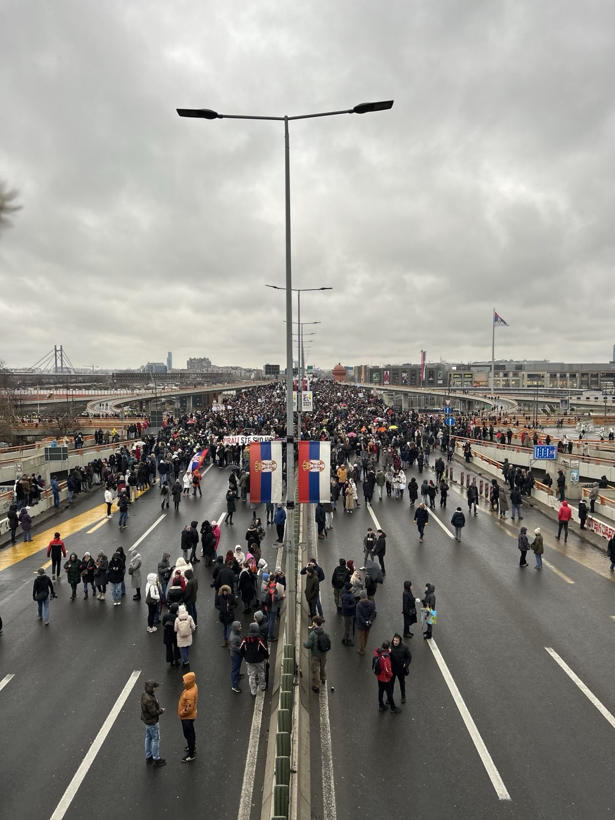 The image shows a large crowd of people gathered on a wide roadway under an overcast sky. The road, divided by metal barriers, appears to be a major thoroughfare in an urban setting. People are walking and standing in groups, many wearing winter jackets, indicating cold weather. In the distance, a bridge spans across the river, and modern buildings line the horizon. Two large light poles are visible in the center foreground, and several national flags are hung along the road. A large structure in the background displays signs featuring images and possible advertisements.