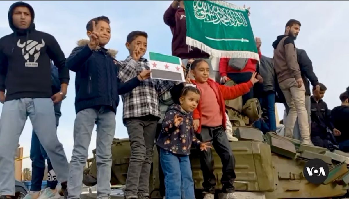 The image depicts a group of people standing on a military vehicle. Central to the image are several children making peace signs, with one holding a flag featuring three red stars on a green, white, and black background. Behind them, other individuals stand, some holding a large green flag with white Arabic script. The sky is clear with a few clouds, creating a bright backdrop. Most people are dressed in casual clothing, including jackets and jeans. In the bottom right corner, a circular logo with "VOA" is visible.