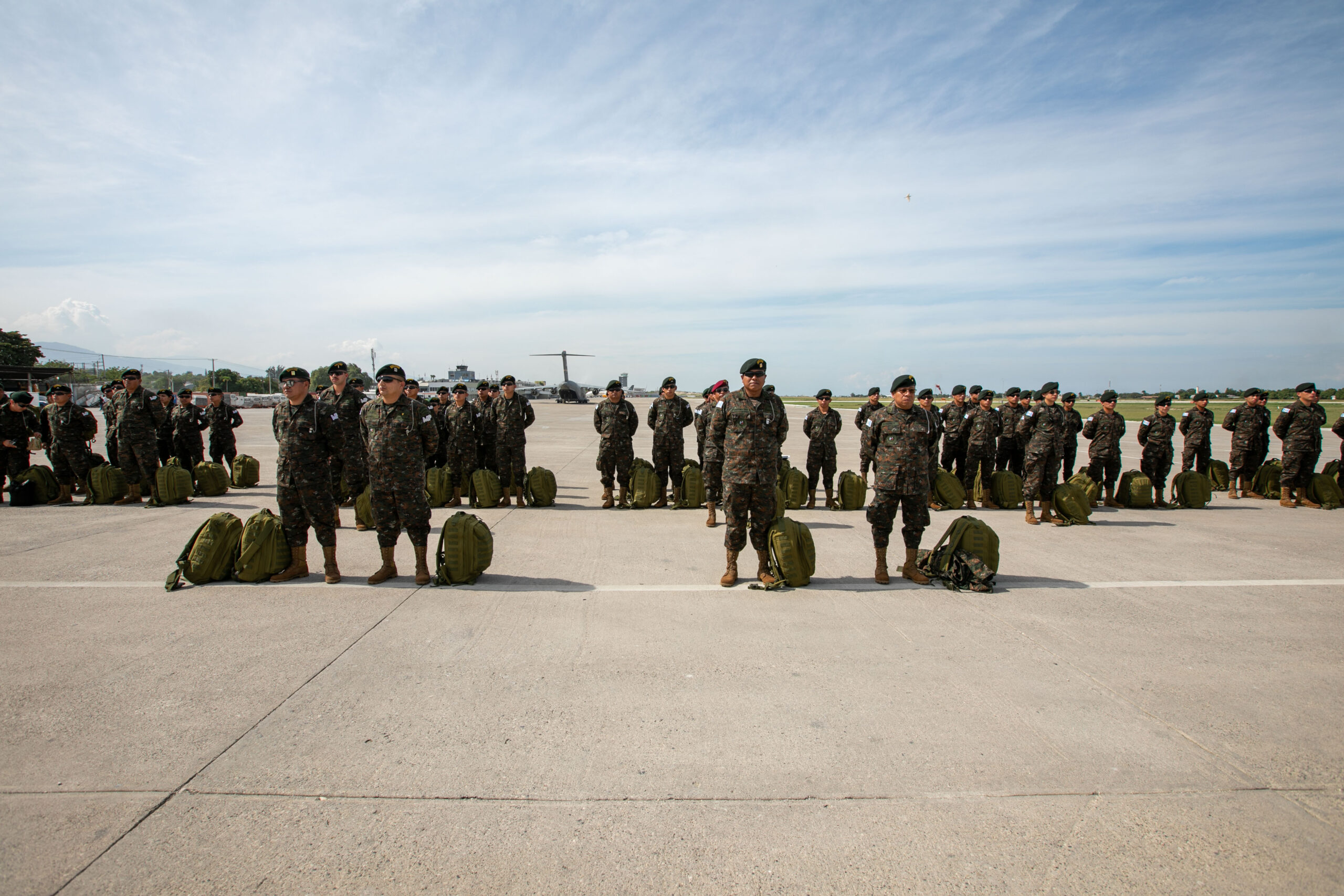 The image depicts a group of military personnel standing in formation on a wide, open tarmac under a clear blue sky with some wispy clouds. The soldiers are dressed in camouflage uniforms and dark berets, and each stands beside a green backpack. The formation consists of several rows of soldiers, extending across the concrete surface. In the background, an airport control tower and a military aircraft, possibly a helicopter, can be seen. The expansive sky above and the distant horizon create a sense of open space.