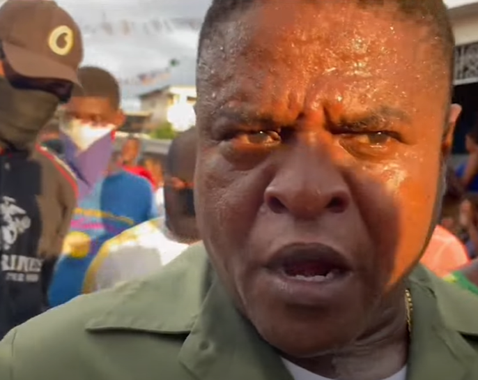 The image shows a close-up of a person wearing a green shirt, appearing to be outdoors. The person's face is glistening, likely due to sweat, and has a serious expression. In the background, there are several other people with various appearances; one person is wearing a hat and a black mask, and another is wearing a white face mask. Some colorful clothing is visible, and the crowd appears blurred due to focus on the foreground subject. The lighting suggests warm, natural sunlight possibly during late afternoon.