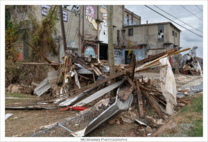 The image shows an abandoned industrial site with significant debris in the foreground. Piles of wooden beams, corrugated metal sheets, and torn fabric are scattered across the area, suggesting destruction or decay. The background features a weathered, multi-story building with peeling paint and overgrown vegetation. Graffiti art, including a pink donut and a few abstract shapes, decorates the walls of the building. Several windows are broken, and the structure appears neglected. The scene is overcast, adding a dreary atmosphere. A fence lies fallen, partially obscured by the debris.