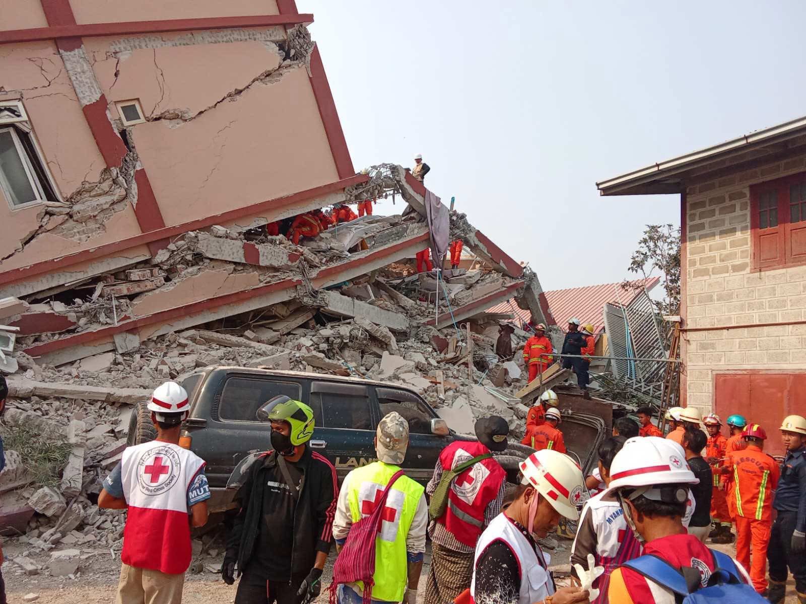 The image depicts a disaster scene with rescue workers amid the rubble of a collapsed building. The multi-story structure shows severe structural damage, with large cracks and areas of concrete debris. Several rescue workers in bright orange uniforms and helmets are searching through the debris, highlighting ongoing rescue efforts. A partially crushed black vehicle is visible among the rubble. In the foreground, people wearing safety gear, including helmets and face masks, are gathered, likely coordinating the rescue. Adjacent to the collapsed building, another structure with red-tinted wooden windows and a tiled roof stands intact.