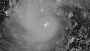 The image shows a satellite view of a hurricane in grayscale, with swirling clouds forming a circular pattern. The eye of the hurricane is clearly visible at the center, appearing as a small circular area with a lighter shade. Around the eye, dense cloud bands spiral outward, creating a textured appearance. The image also features three bluish-white light spots near the top center, likely indicating lightning or other atmospheric activity. On the right side, scattered clouds vary in density and form intricate patterns against a darker background.