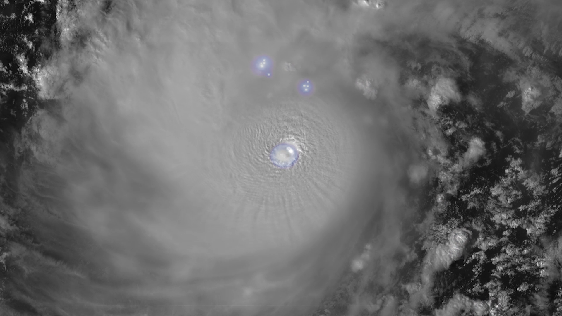 The image shows a satellite view of a hurricane in grayscale, with swirling clouds forming a circular pattern. The eye of the hurricane is clearly visible at the center, appearing as a small circular area with a lighter shade. Around the eye, dense cloud bands spiral outward, creating a textured appearance. The image also features three bluish-white light spots near the top center, likely indicating lightning or other atmospheric activity. On the right side, scattered clouds vary in density and form intricate patterns against a darker background.