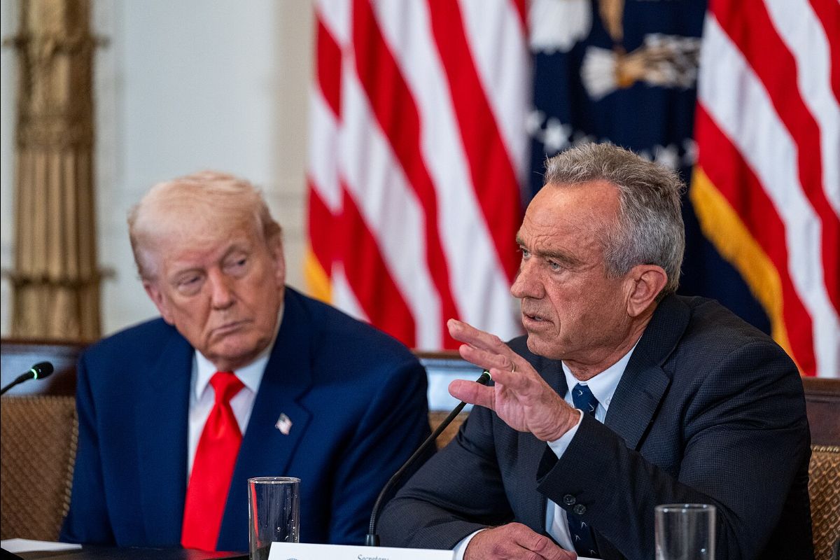 The image depicts two men seated at a table with a backdrop of American flags. The man on the left wears a dark suit with a white shirt and a red tie. His hair is light and he is looking towards the man on the right. The man on the right is also in a dark suit, with a white shirt and a dark tie. He gestures with his right hand as if emphasizing a point. There are microphones in front of each of them and a blurred piece of paper is visible in the foreground.