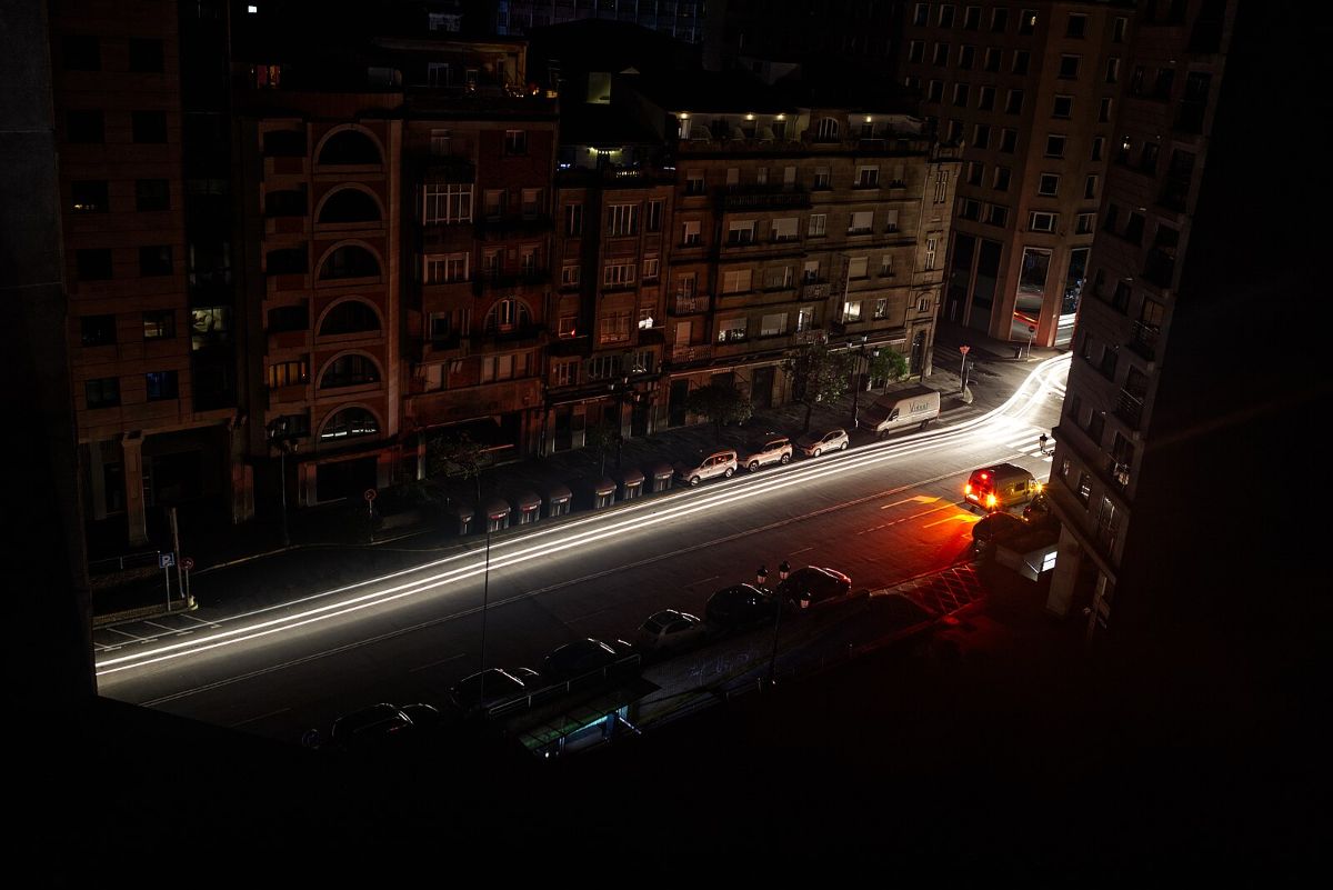 The image depicts an urban nighttime scene with a long exposure photograph of a street. The street, filled with light trails from passing vehicles, runs horizontally across the image. The light trails are mostly white, suggesting headlights, with a bright red spot on the right indicating a vehicle's taillights or brake lights. Buildings frame the street on either side, with multiple stories visible. The structures are dark, with only a few windows softly illuminated, hinting at minimal activity. The foreground building has arched architectural features and balconies. Parked cars line the street, casting long shadows due to the streetlights and vehicle lights.