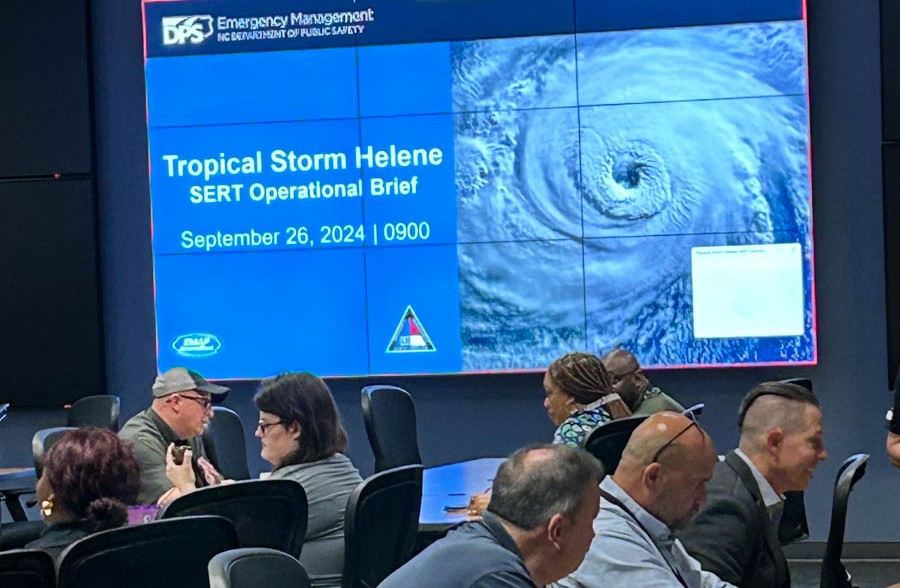 The image depicts a conference room where a group of people is seated at tables. They are engaged in discussions or listening intently, with several individuals typing on laptops. The focus is on a large digital screen displaying an emergency management briefing. The screen is divided into several sections; the left shows text on a blue background while the right displays a satellite image of a swirling storm. The setting appears to be a formal meeting or briefing related to a tropical storm.