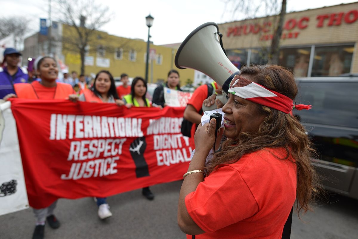 The image depicts a street protest where several people are participating. In the foreground, a woman wearing a red bandana and red shirt holds a megaphone, passionately speaking into it. Behind her, a group of people march, holding a large red banner. The banner has bold white text and an image of a raised fist in black. The participants are wearing various colored shirts and jackets, with some in orange safety vests. The background shows blurred buildings and a tree, creating an urban setting. The scene conveys a sense of activism and solidarity.