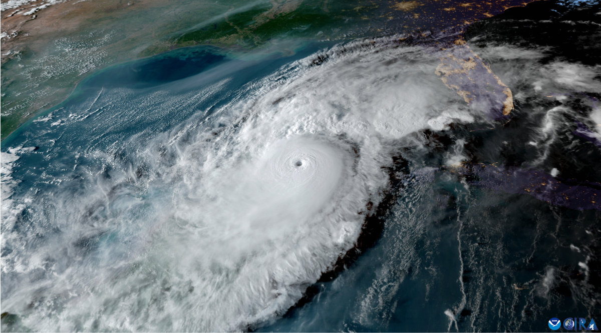 The image is a satellite view of a massive hurricane over the ocean. At the center is the eye of the hurricane, a well-defined circular area surrounded by tightly wound, swirling clouds. The hurricane spans most of the visible section of the ocean, with dense, white cloud formations spiraling outward. At the top, there is a view of the mainland, where patches of green and brown land are visible. The ocean beneath the clouds is deep blue, with lighter patches indicating waves or disturbed water. The right side of the image is darker, possibly approaching nightfall or an area with less cloud cover, revealing some city lights on the mainland.