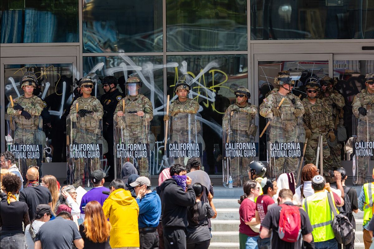 The image depicts a group of California National Guard soldiers standing in a line outside a building with large glass doors. They are wearing camouflage uniforms and helmets, and each holds a transparent shield with “California National Guard” printed on it in white letters. The doors behind them are marked with graffiti in various colors. In front of the soldiers, a crowd of civilians is gathered, some wearing masks and bright-colored clothing. The scene suggests a protest or public gathering, with people interacting or observing the line of guards.