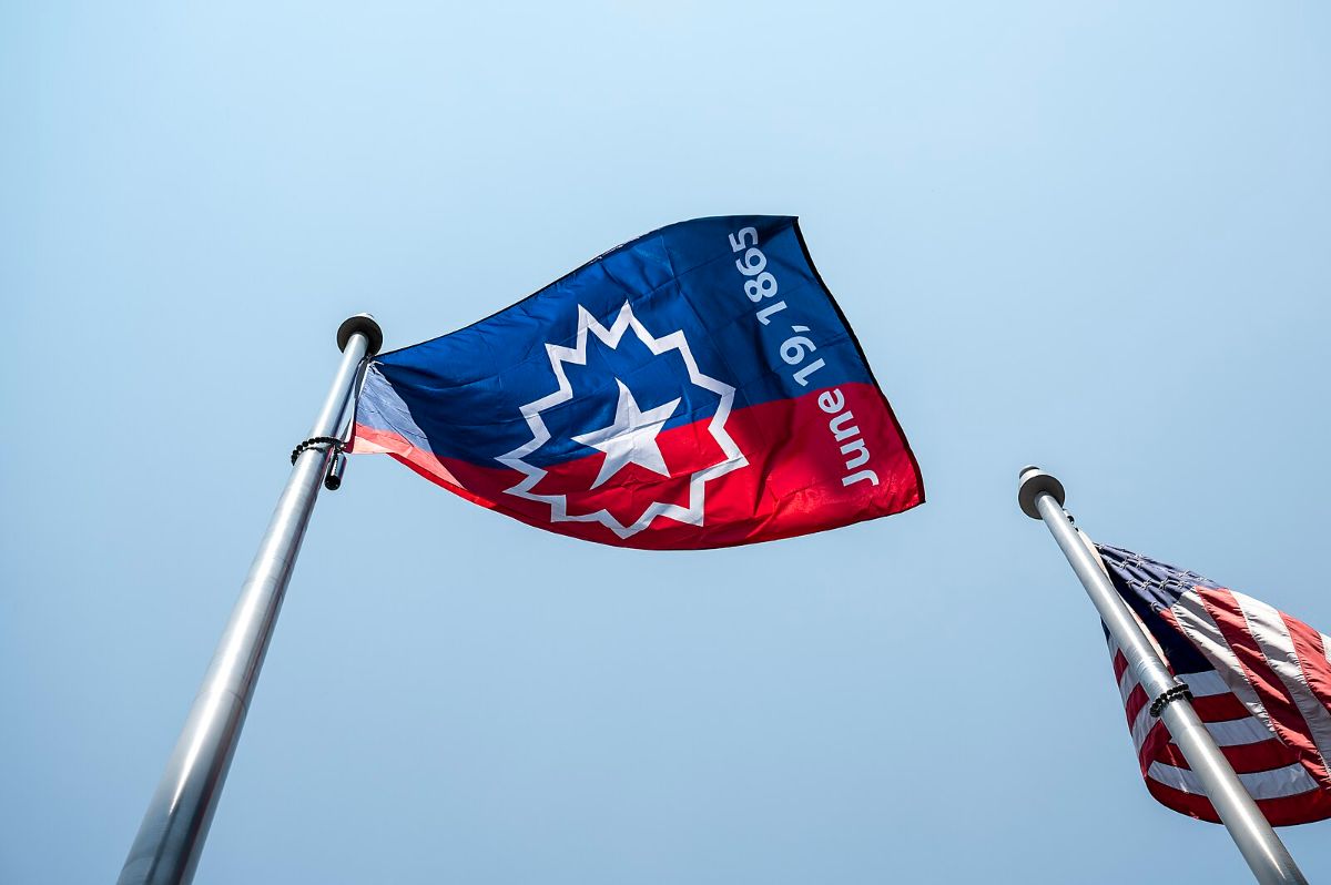 The image shows two flags raised on flagpoles against a clear blue sky. The central flag, prominently displayed, features a design with a blue field on the left, transitioning to red on the right, and is bisected by a white star surrounded by a burst pattern. The text "June 19, 1865" is printed vertically along the blue section. To the right, partially visible, is an American flag with its familiar red and white stripes and blue canton with white stars. The flagpoles are metallic and extend upwards at a slight angle, emphasizing the flags against the sky.