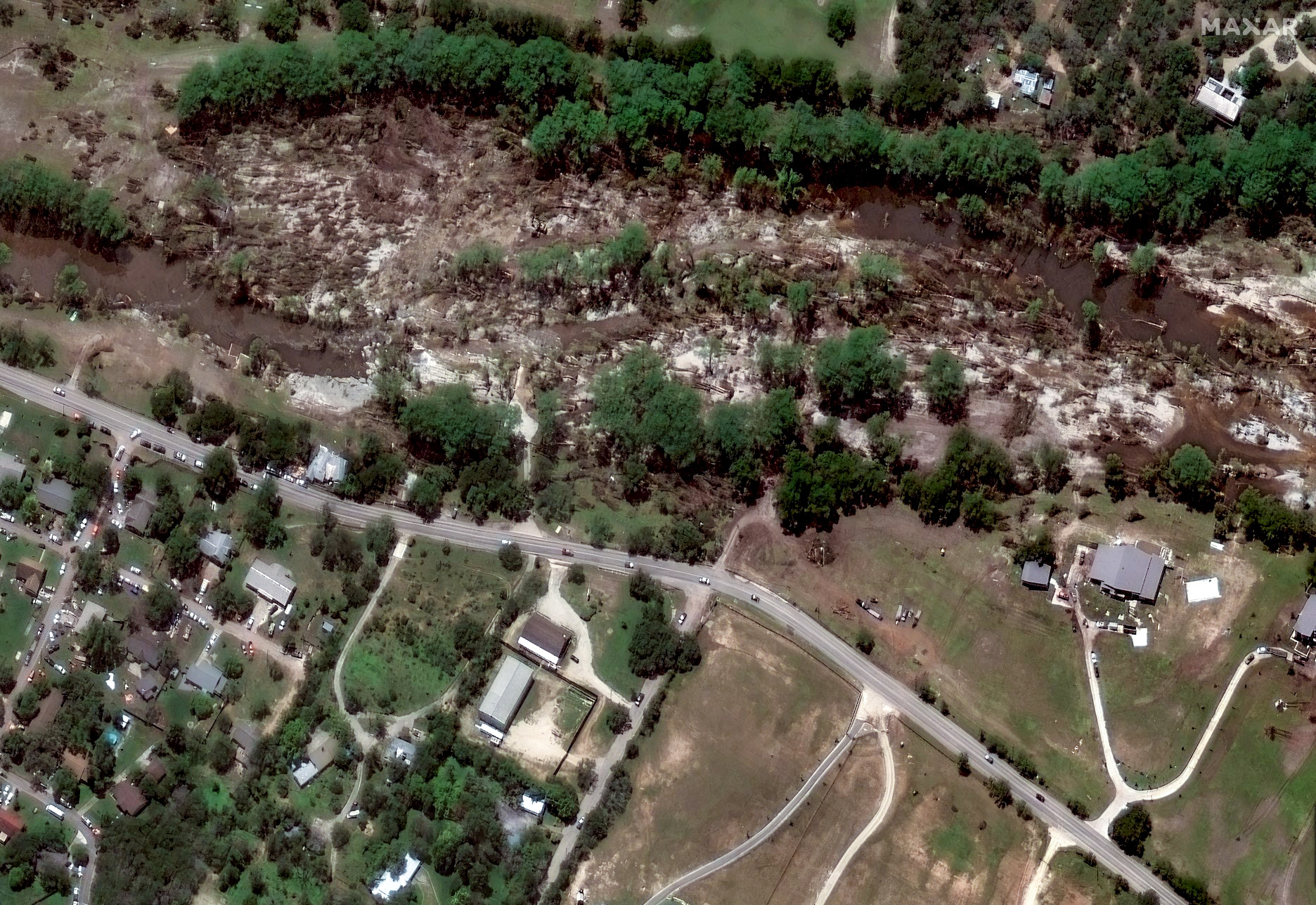 The image is a satellite view of a landscape depicting a river surrounded by dense green foliage. The river flows horizontally, cutting through a wooded area in the upper part of the image. The landscape surrounding the river shows signs of uneven terrain with patches of brown and exposed earth, suggesting flooding or erosion. Below the river, there is a road running parallel to it, with several houses and a building complex on either side. The houses are organized into a small residential area with a variety of smaller structures and green lawns, typical of a suburban setting. A few cars are visible on the road, indicating some level of activity. Further south, the landscape becomes more open, featuring fewer trees and larger patches of grassland.