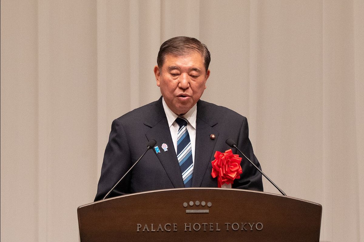 The image shows a man standing behind a lectern, delivering a speech. He is wearing a dark suit with a white dress shirt and a striped tie. A red ribbon is pinned to his suit jacket. The lectern is simple and dark-colored with microphones placed on it. The background is plain with vertical lines, suggesting a formal or official setting. The lighting is soft, focusing primarily on the man.
