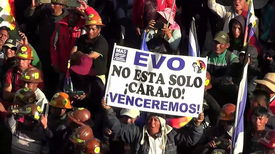 The image shows a large group of people gathered outdoors, many wearing hard hats, which appear to be miners' helmets. The crowd is dense and fills the frame, with individuals dressed in various colors, including red, green, and brown. Some people are holding flags, and there is a prominent white sign in the center with bold blue text held by a person wearing a hat and a jacket. The atmosphere suggests a public demonstration or rally.
