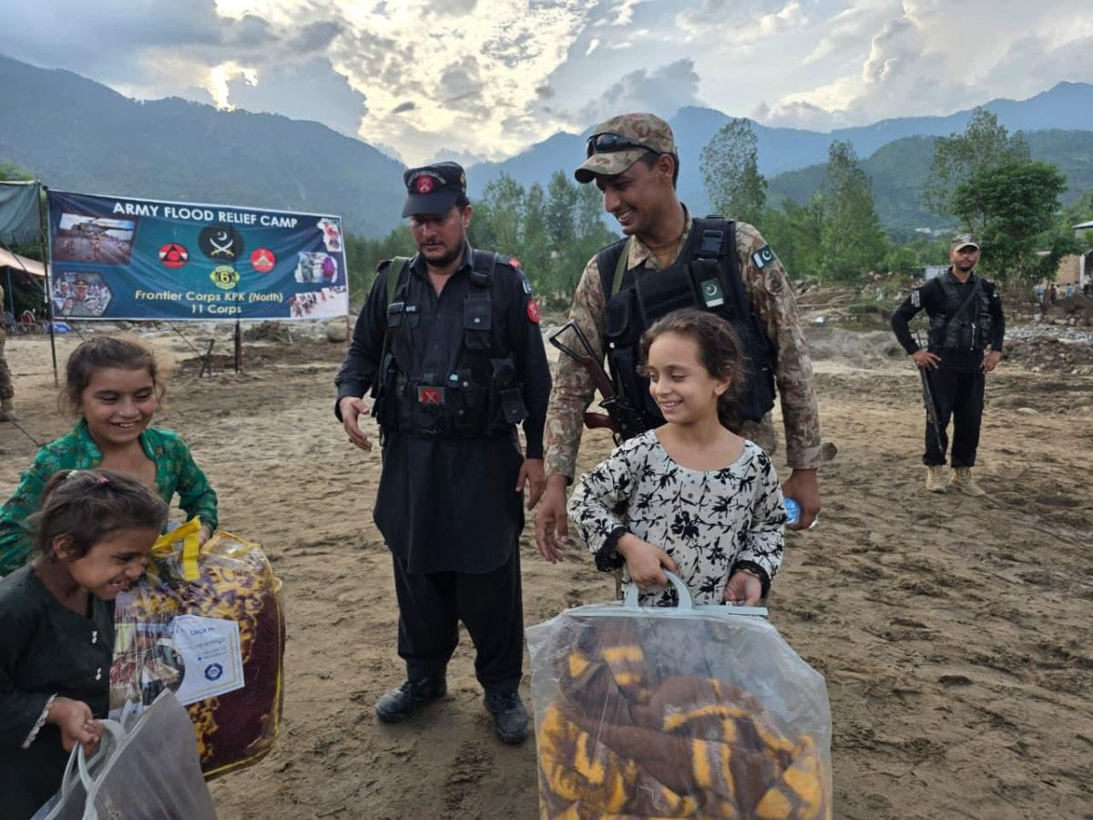 The image depicts a relief camp scene in a mountainous area, under a cloudy sky. In the foreground, three children are smiling and carrying plastic bags filled with colorful blankets. Two uniformed military personnel are accompanying them; one is in dark uniform, and the other in camouflage. Both have arm patches with a flag design. In the background, the landscape features mountains and trees. There is a large banner that reads "Army Flood Relief Camp" alongside other text, displaying various emblems and logos. The ground appears muddy, suggesting recent flooding.