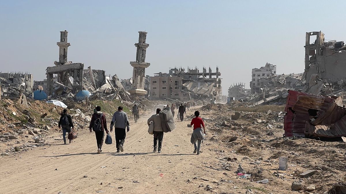 The image depicts a scene of destruction, with several people walking along a dirt road flanked by debris and ruined buildings. In the foreground, a group of individuals, including a person in a red shirt, walk towards the background. The path is surrounded by rubble, with remnants of structures on both sides. Two tall, partially-collapsed buildings stand prominently to the left and right, with their upper sections leaning at precarious angles. In the distance, more damaged buildings are visible, highlighting extensive structural damage. The sky is clear, providing stark contrast to the scene of devastation below.