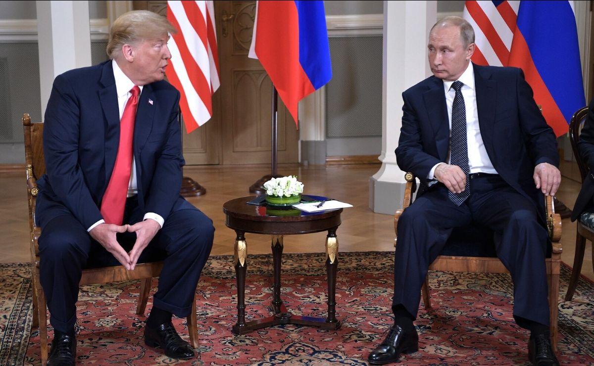 The image shows two men formally dressed and seated on wooden chairs with plush cushions. They are positioned on either side of a small round table that holds a floral arrangement and some papers. Behind them, the background reveals a few standing flags with red, white, and blue colors. The floor is adorned with a red patterned rug, and the setting appears to be indoors with wooden doors and ornate molding visible. Both men seem to be engaged in a serious conversation, with one gesturing slightly with his hands.