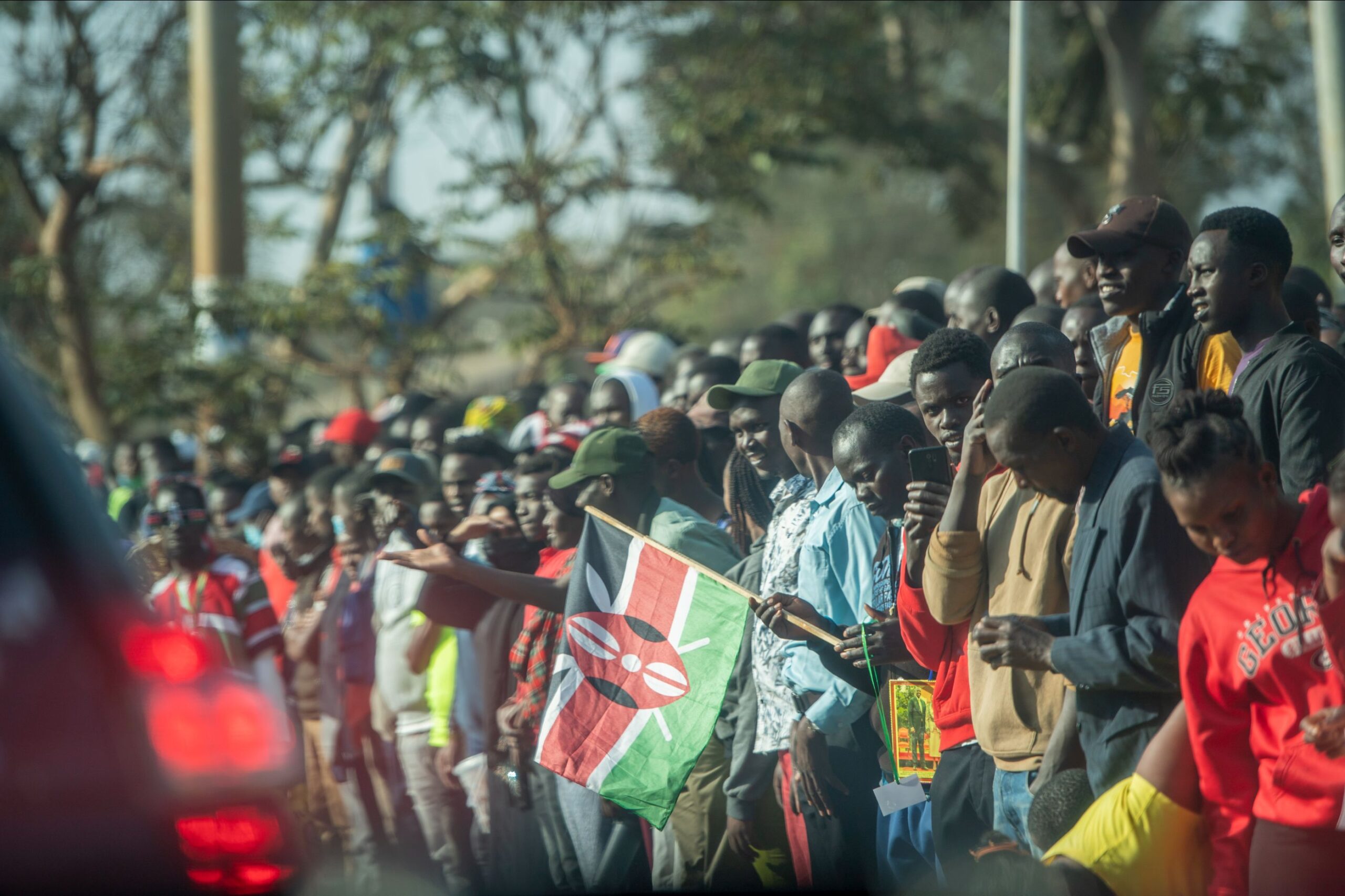 The image depicts a crowd of people standing closely together on what appears to be the side of a street. Many individuals in the crowd are wearing casual clothing, and some are dressed in bright colors, like red and yellow. A number of people are holding up mobile phones, possibly recording or photographing something off-frame. One individual is prominently holding a Kenyan flag. The setting seems to be outdoors with blurred trees and a pole in the background. The lighting suggests it might be a sunny day. In the foreground, the blurred rear lights of a vehicle are visible, glowing red.