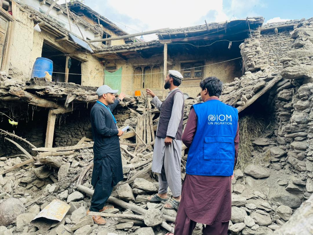 The image shows three men standing amidst the rubble of a damaged building. The structure appears to be made of wood and stones, with parts of the walls and roof collapsed or damaged. A blue barrel is visible on what remains of the upper level. One man, wearing traditional dress with a cap, gestures towards the building while talking to another man. The third man wears a bright blue vest with the IOM logo, indicating a possible connection to the International Organization for Migration. The setting is outdoors, with a partly cloudy sky above.