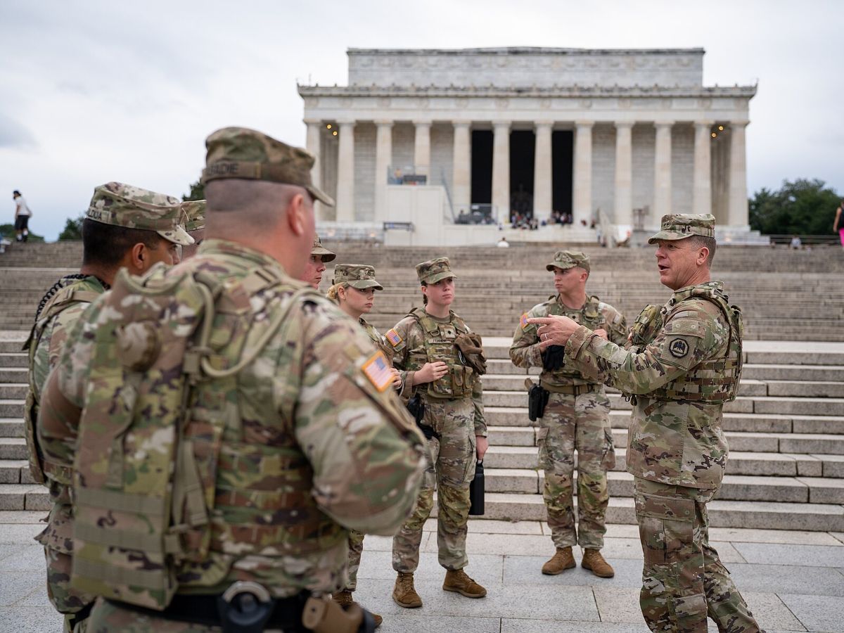The image depicts a group of military personnel dressed in camouflage uniforms gathered on the steps of a large, neoclassical building. The building in the background is the Lincoln Memorial, identifiable by its iconic columns and central statue visible through the columns. The sky is overcast, casting a soft light over the scene. In the foreground, several soldiers face each other, engaged in conversation. They wear helmets and tactical gear. The soldiers' attire blends in with the grayscale tones of the stone steps. A few people can be seen in the background on the steps, adding a sense of depth and scale to the scene.