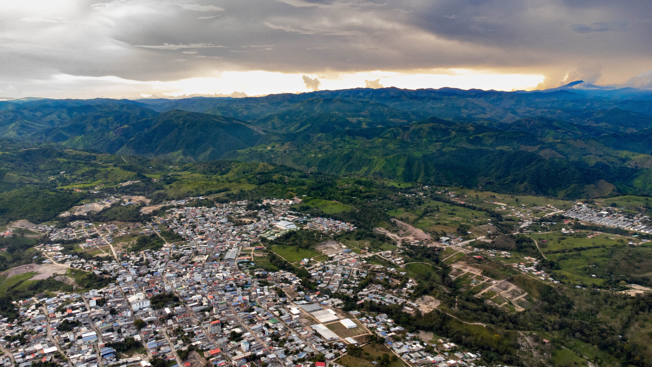 The image is an aerial view of a sprawling town surrounded by lush, green mountainous terrain. In the foreground, numerous buildings and streets form a dense urban area with a mix of residential and commercial structures. Roofs vary in color, adding a patchwork effect. Beyond the town, expansive rolling hills and mountains dominate the landscape, covered with dense green foliage. The sky above is overcast with thick gray clouds, but a streak of brighter light peeks through near the horizon, creating a contrast between the darkened sky and the illuminated landscape.