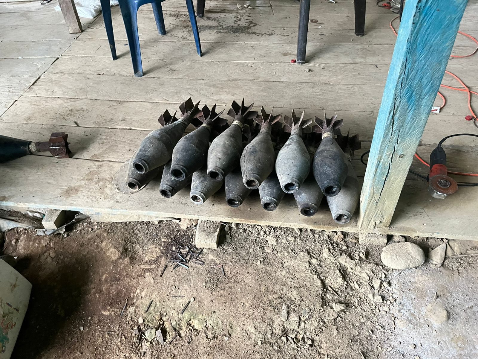 The image shows a collection of mortar shells laid out on a wooden surface. There are nine shells arranged in two rows, with the pointed tips facing the camera. Each shell has a dark, metallic body and fins at the rear. The wooden floor appears aged with visible dirt and wear marks. To the right, there is a blue wooden post and an orange power tool with a disc attached, sitting on the floor. In the background, part of a blue plastic chair and wooden planks can be seen, along with some scattered nails on the dirt floor beneath the wooden surface.
