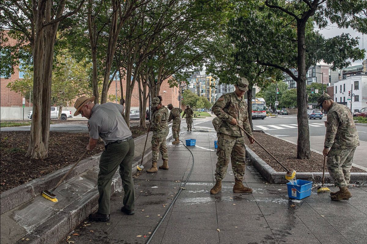 The image depicts a group of seven individuals, dressed in military camouflage uniforms and one in a park ranger outfit, cleaning a concrete sidewalk in a city environment. They are holding long-handled brushes, scrubbing the surface which appears wet and soapy, suggesting they are engaged in cleaning or maintenance work. The setting is an urban sidewalk lined with trees, providing shade. In the background, there are buildings, including a brick structure and modern apartments. A city bus and several cars are visible on the adjacent street.