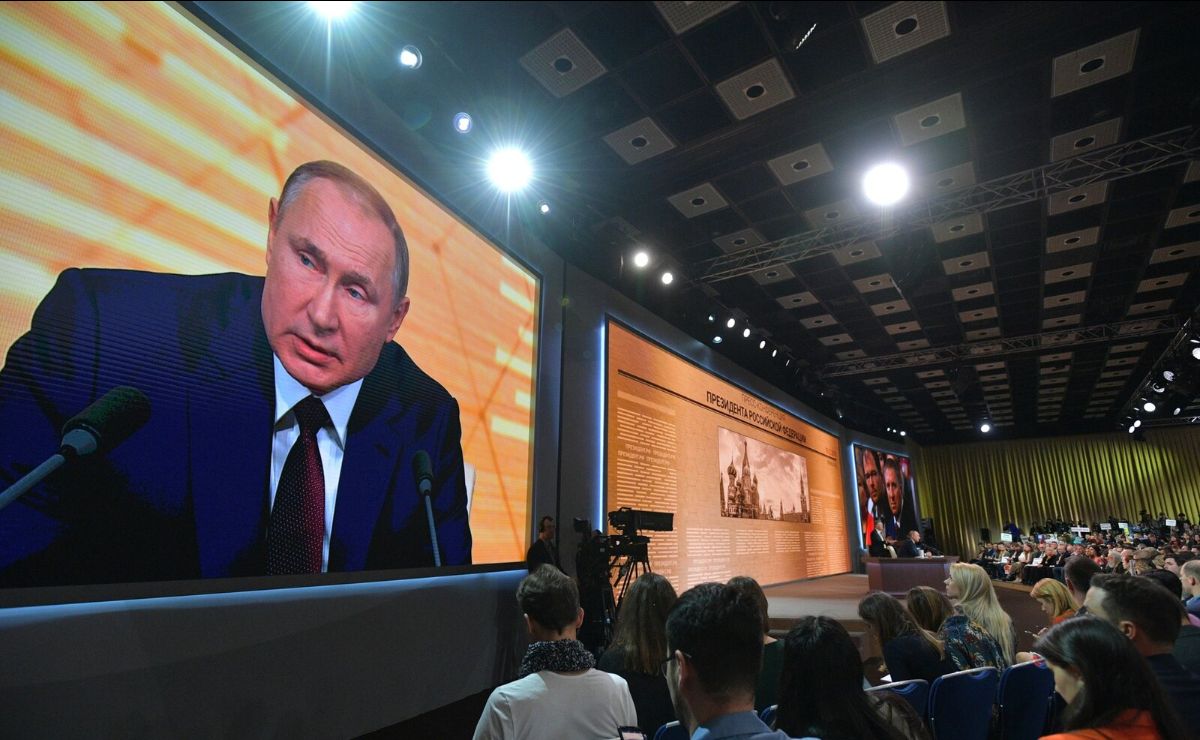 The image shows a large conference room with an audience seated facing a stage. On the left side of the image, a large screen displays the image of a person wearing a dark suit and tie, speaking into a microphone. The room has a patterned ceiling with lights illuminating the stage area. On the right side, there is a long panel with text and an image of a building. The audience consists of many people sitting closely together, observing the presentation.