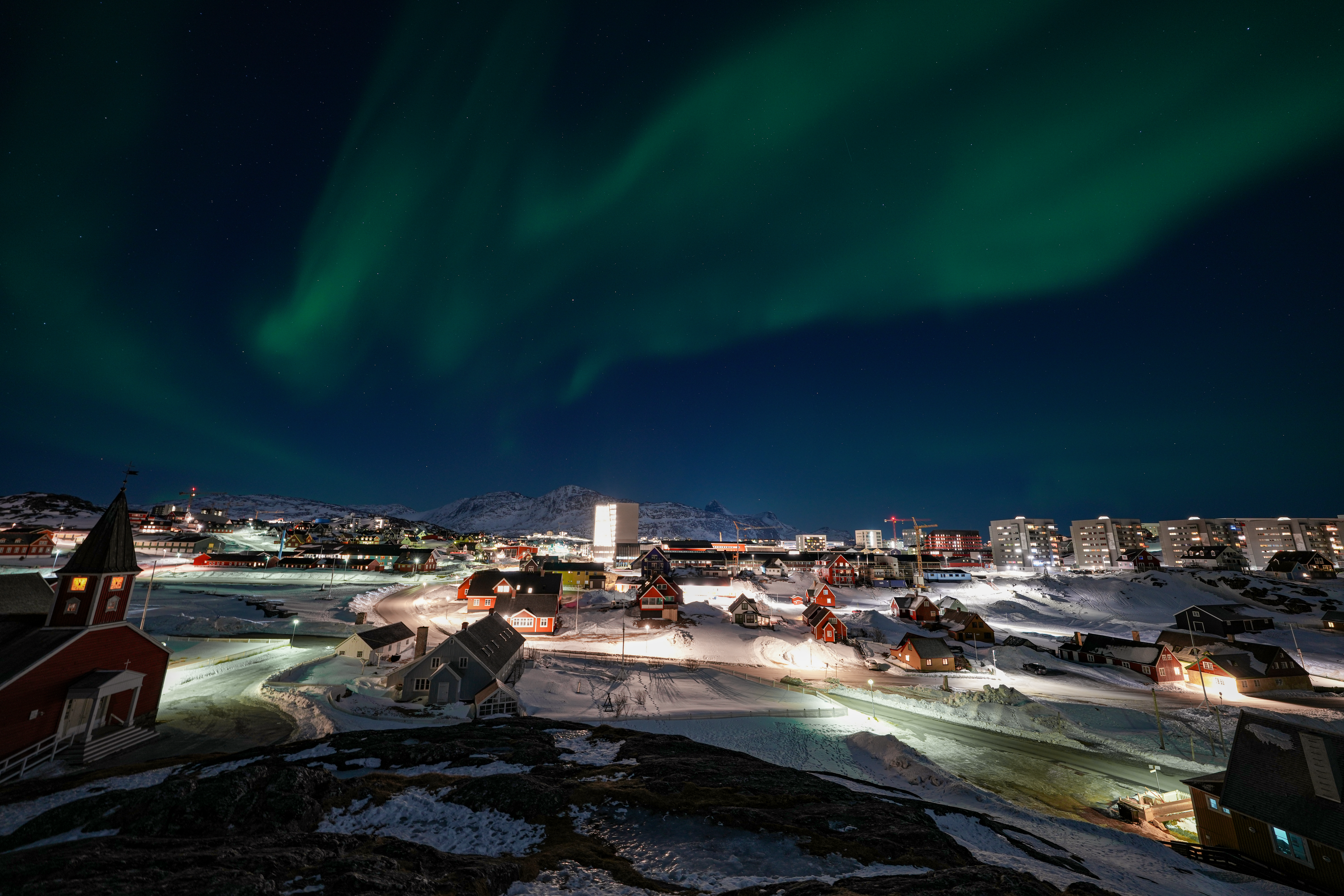 The image depicts a nighttime scene in a snowy, urban area illuminated by artificial lights. In the sky, vibrant green auroras stretch and swirl against a backdrop of stars. The foreground features a church with a pointed roof and red siding, alongside various residential buildings and roads blanketed in snow. In the background, modern apartment buildings are visible, and snow-covered mountains rise in the distance. The cityscape is bathed in a cold, bluish light, contrasting with the warm glow from the buildings and streetlights.