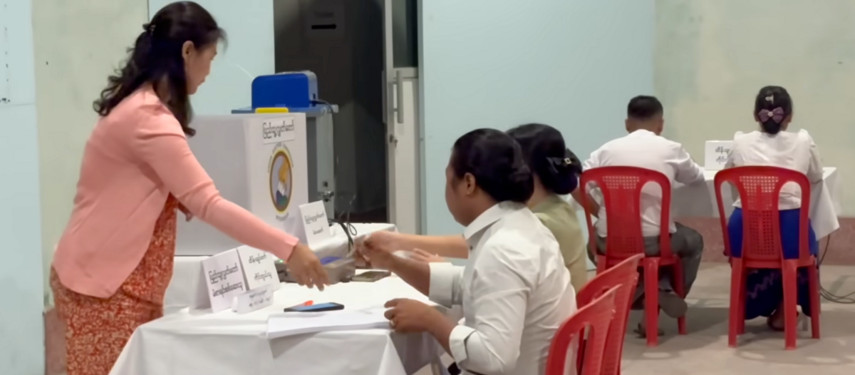 A woman handing over a paper at a voting table with a ballot box nearby.
