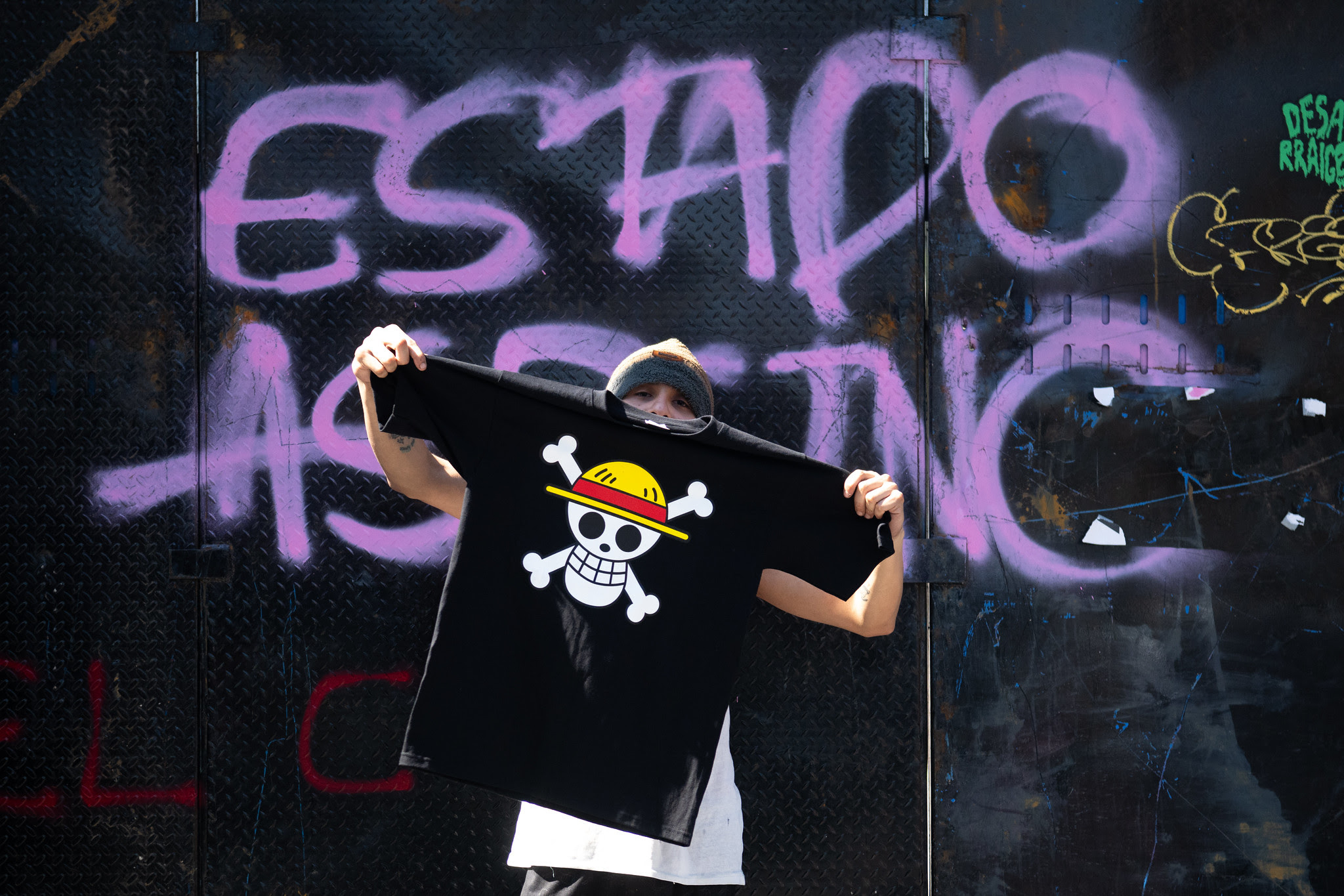 Person holding a T-shirt with a cartoon skull design in front of a wall with graffiti.
