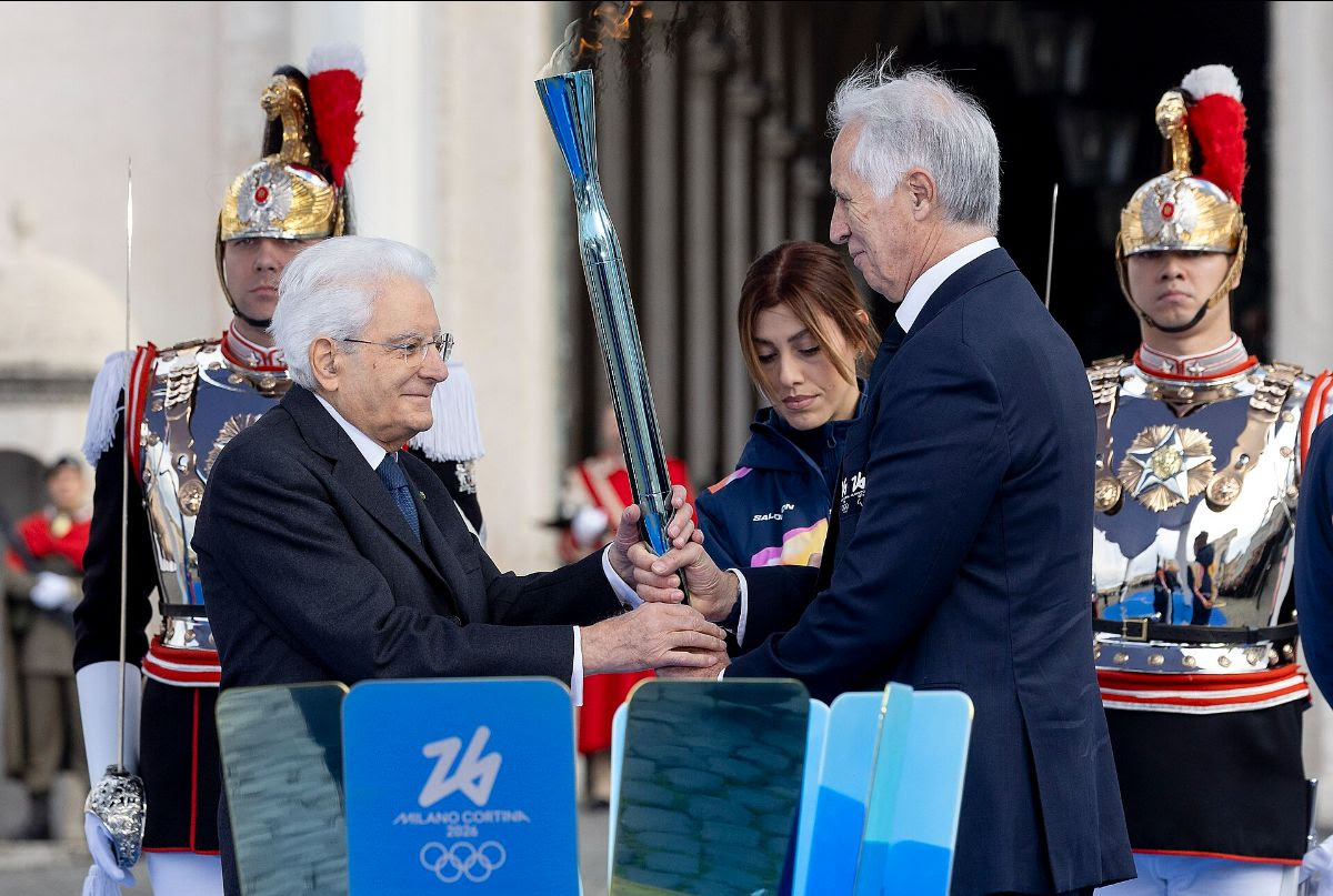 Two men exchange a blue torch during a formal ceremony, flanked by two uniformed guards.