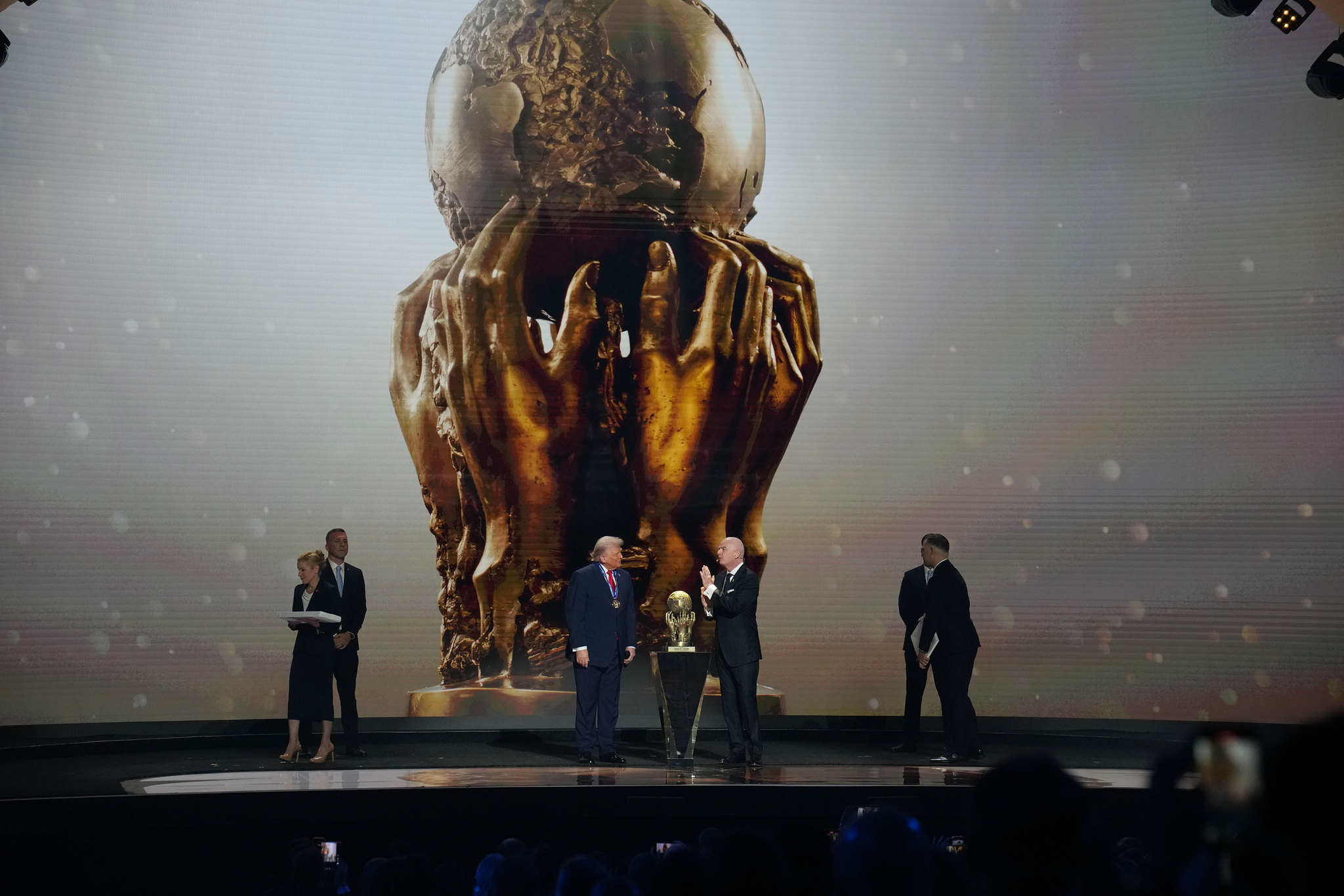 The image shows a formal event on a stage featuring a large backdrop displaying an enormous depiction of a trophy. The trophy consists of a globe held by two large, intricately detailed gold hands. In the foreground, on the stage, three people in suits and a woman holding a stack of papers are present. The scene is lit with dramatic stage lighting, casting shadows and creating a focused atmosphere. A small replica of the trophy is placed on a small, clear stand at the center of the stage.