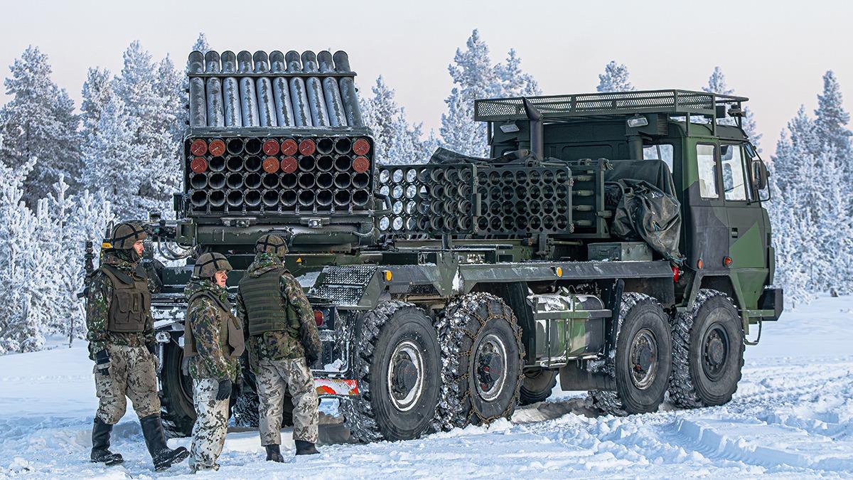 The image shows a large military vehicle equipped with a rocket launcher system in a snowy forest setting. The vehicle is painted in a camouflage pattern and has a robust, multi-wheeled design. The rear section features multiple cylindrical tubes arranged in rows, characteristic of a rocket launcher. Three soldiers, dressed in winter camouflage gear and helmets, stand beside the vehicle, appearing to converse. The ground is covered in snow, and the surrounding landscape is filled with snow-laden trees under a pale sky.